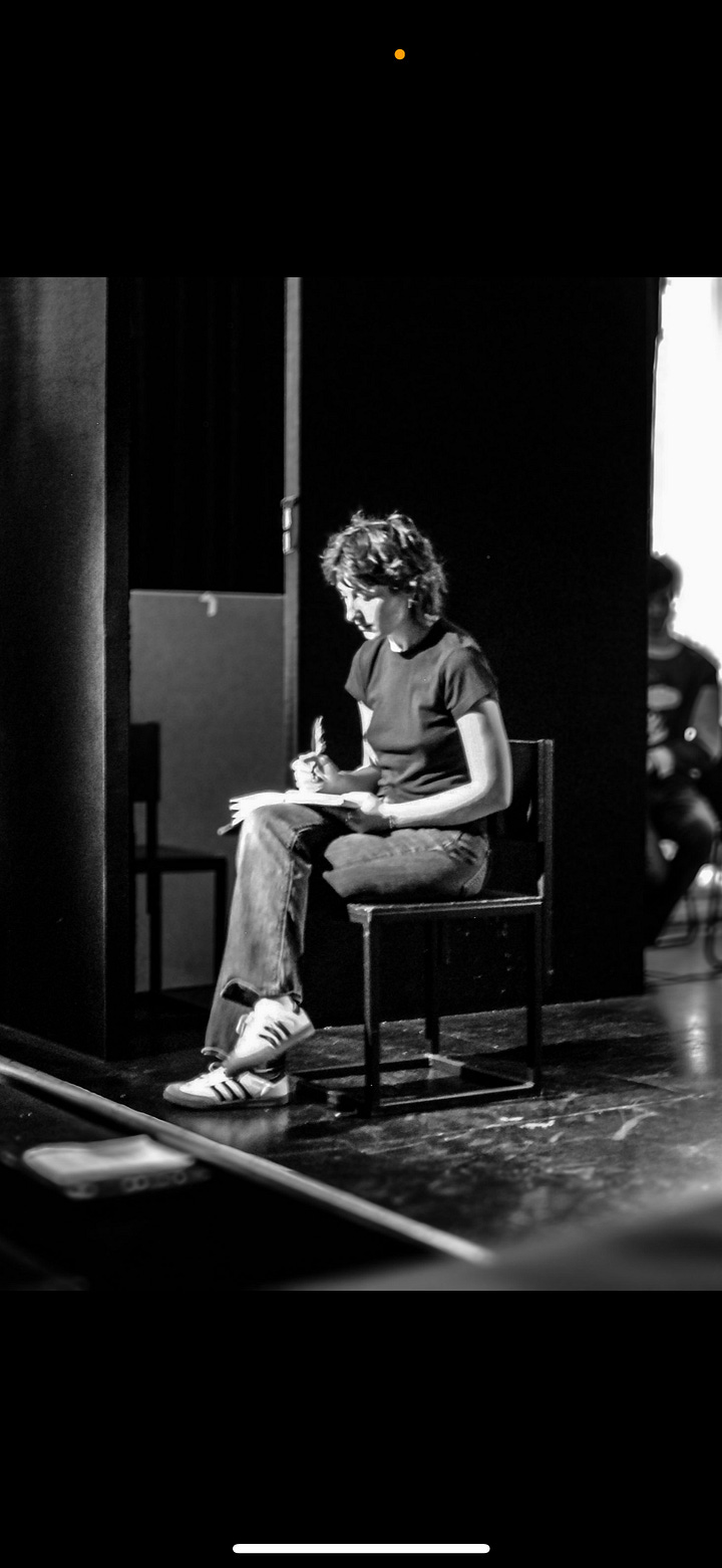 A person with short, tousled brown hair sits on the edge of a stage under dramatic lighting. They wear a plain black t-shirt and jeans, gazing pensively toward a light source. The scene is dimly lit with a deep blue and purple backdrop, and another person stands behind them, visible only from the waist down, wearing jeans with a large knee tear. The atmosphere suggests a theatrical or emotional performance moment. On the rightA black and white photograph of a person seated on a chair at the edge of a stage, focused intently on writing in a notebook balanced on their lap. They wear a dark t-shirt, jeans, and Adidas sneakers. Soft light falls on their face and hands, creating a contemplative atmosphere. In the blurred background, another figure sits quietly in shadow, suggesting a behind-the-scenes or rehearsal setting in a theater.