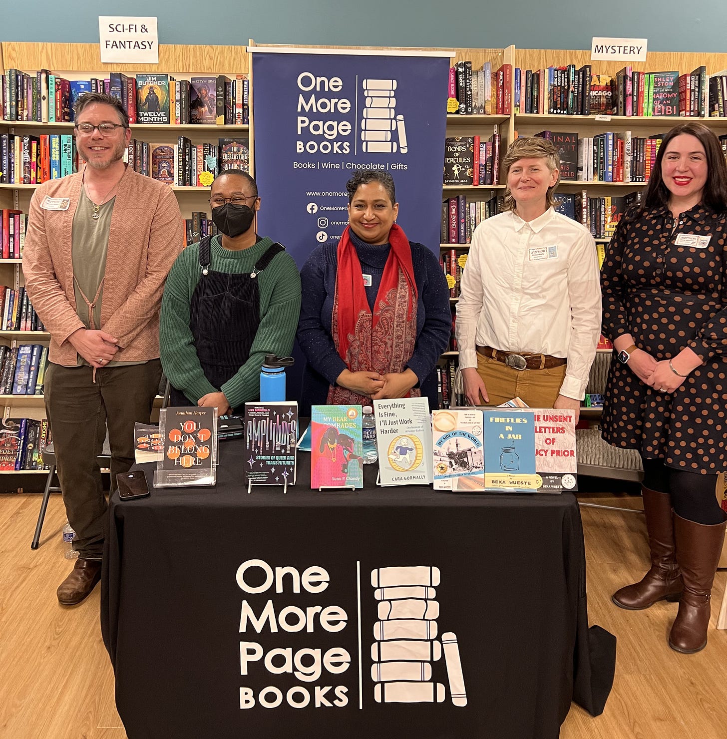 Five authors standing in front of One More Page Books poster