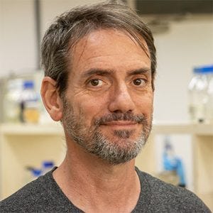 20260727_benyspira homem com olhos escuros, barba curta grisalha e cabelos lisos grisalhos. Usa uma camiseta cinza e está sorrindo levemente com vidrarias de laboratório ao fundo.
