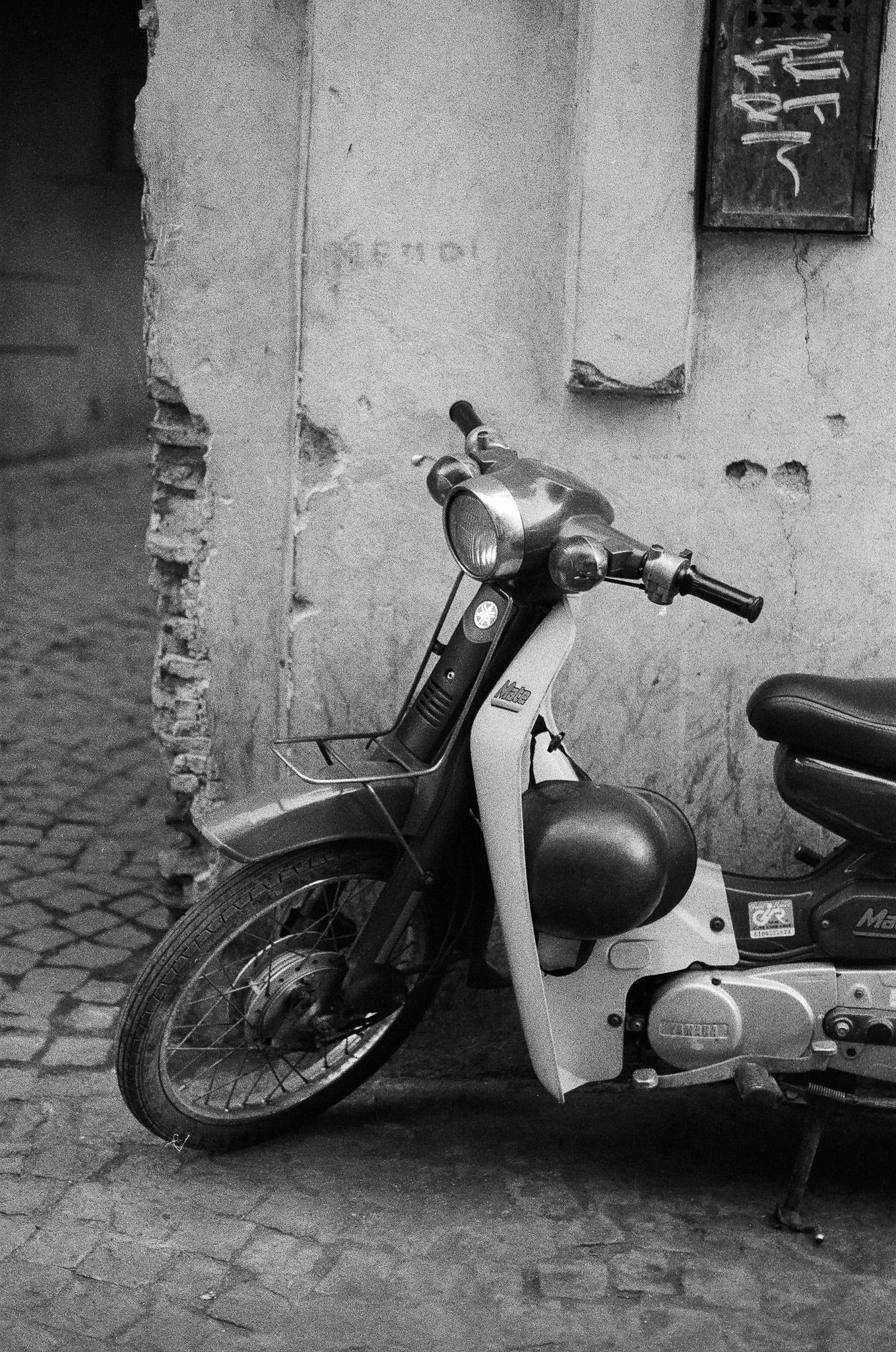 Black-and-white photograph of an old motorbike parked against a textured wall in the Marrakesh Medina, showing worn paint, exposed brick, and cobblestone ground.