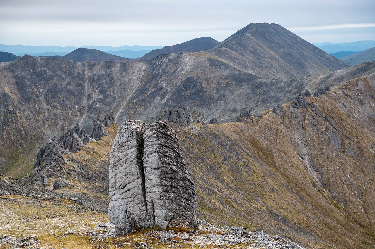 A tall split granite pillar stands in the foreground on a tundra ridge, weathered into two leaning columns, with more jagged tors and a pyramid-shaped peak beyond under a soft grey sky.