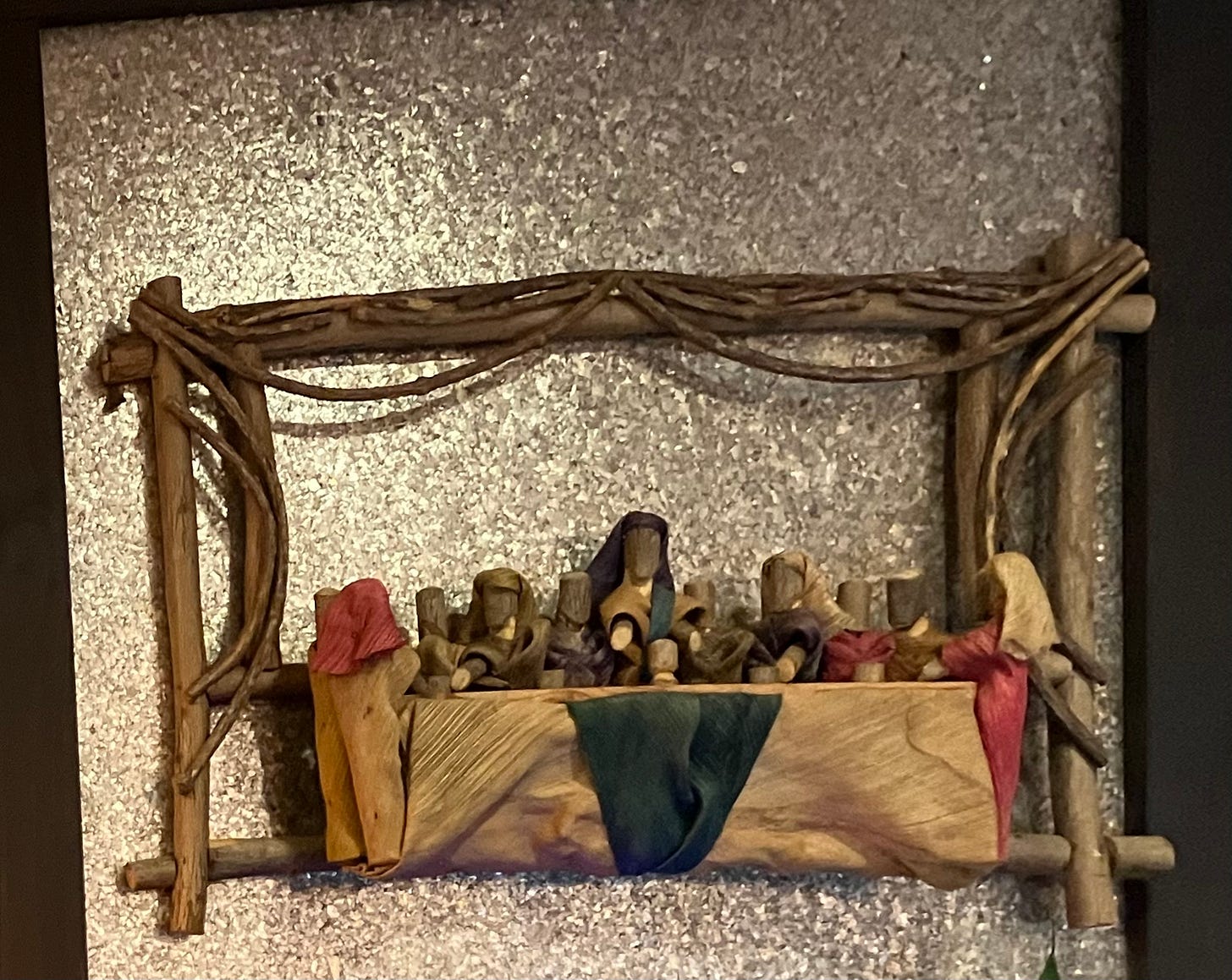 A sculpture made with branches, wood, and leaves depicitnng the last supper. It is set against a shiny silver background in a black frame.