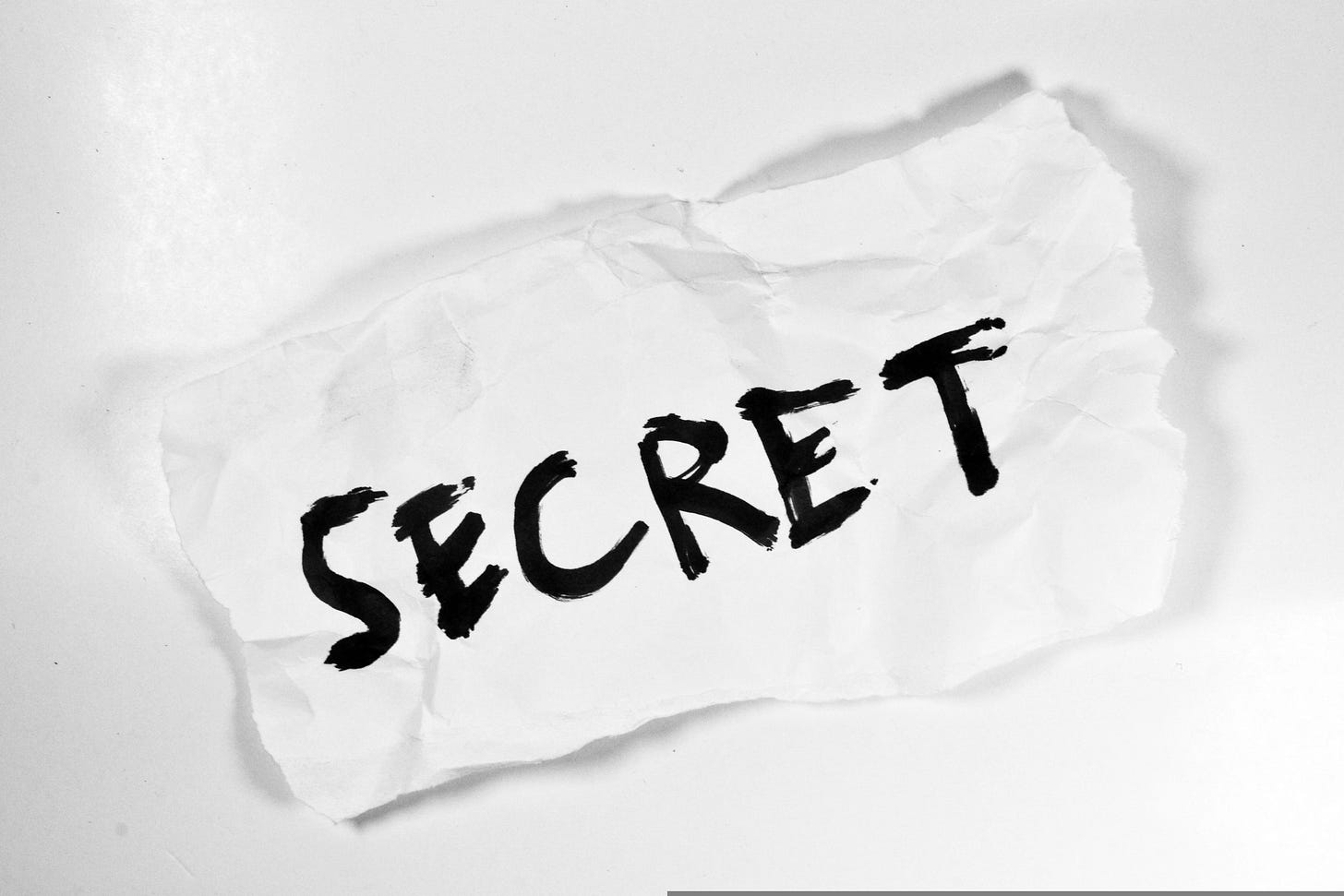 A white crumpled piece of paper rests on a white backgroung. On the paper, the word SECRET is written in thick, black, capital letters. A white crumpled piece of paper rests on a white backgroung. On the paper, the word SECRET is written in thick, black, capital letters.