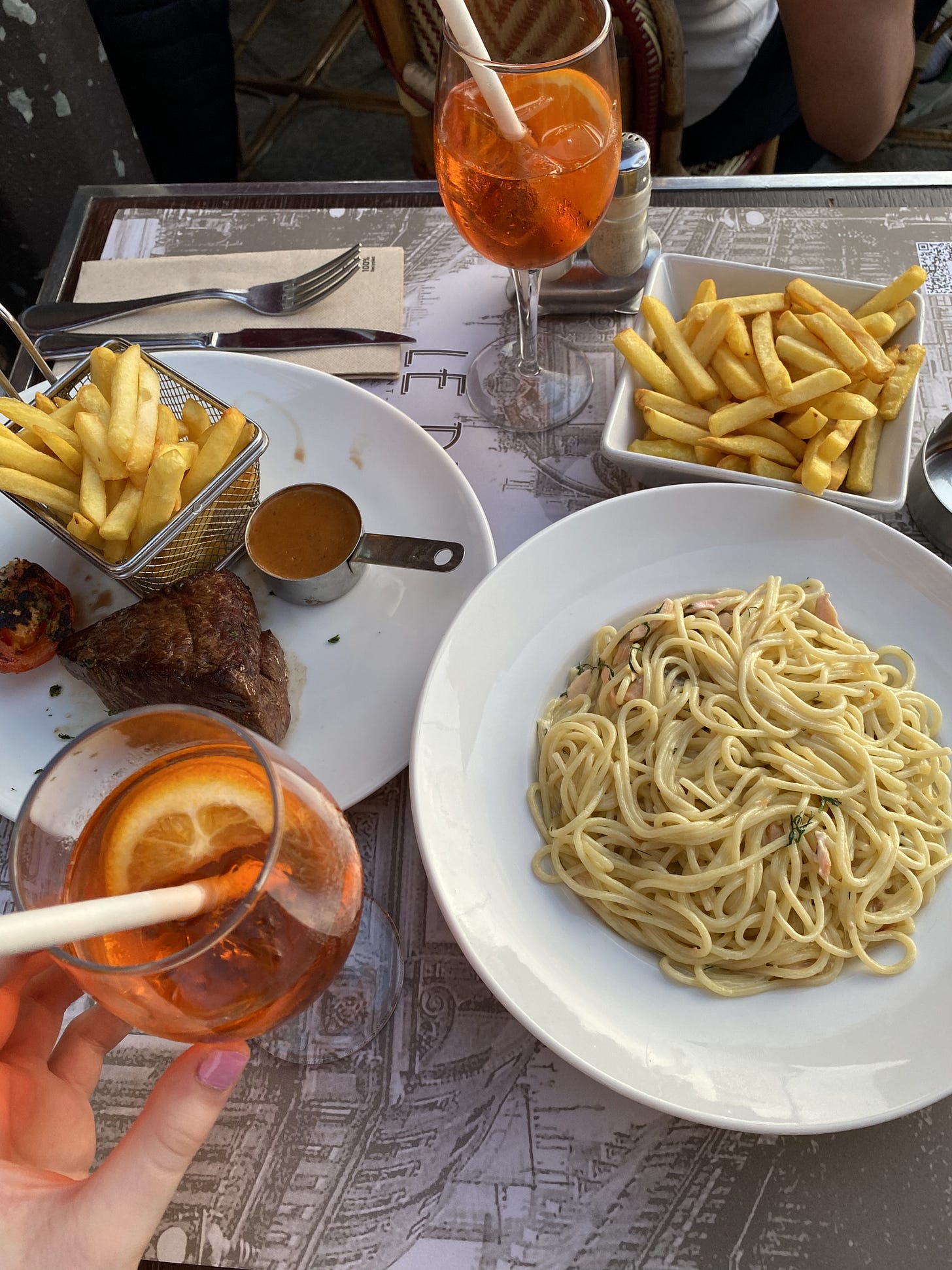 An outdoor table with a bowl of spaghetti, a plate with steak and chips, a bowl of chips, two Aperol Spritzes, and cutlery.