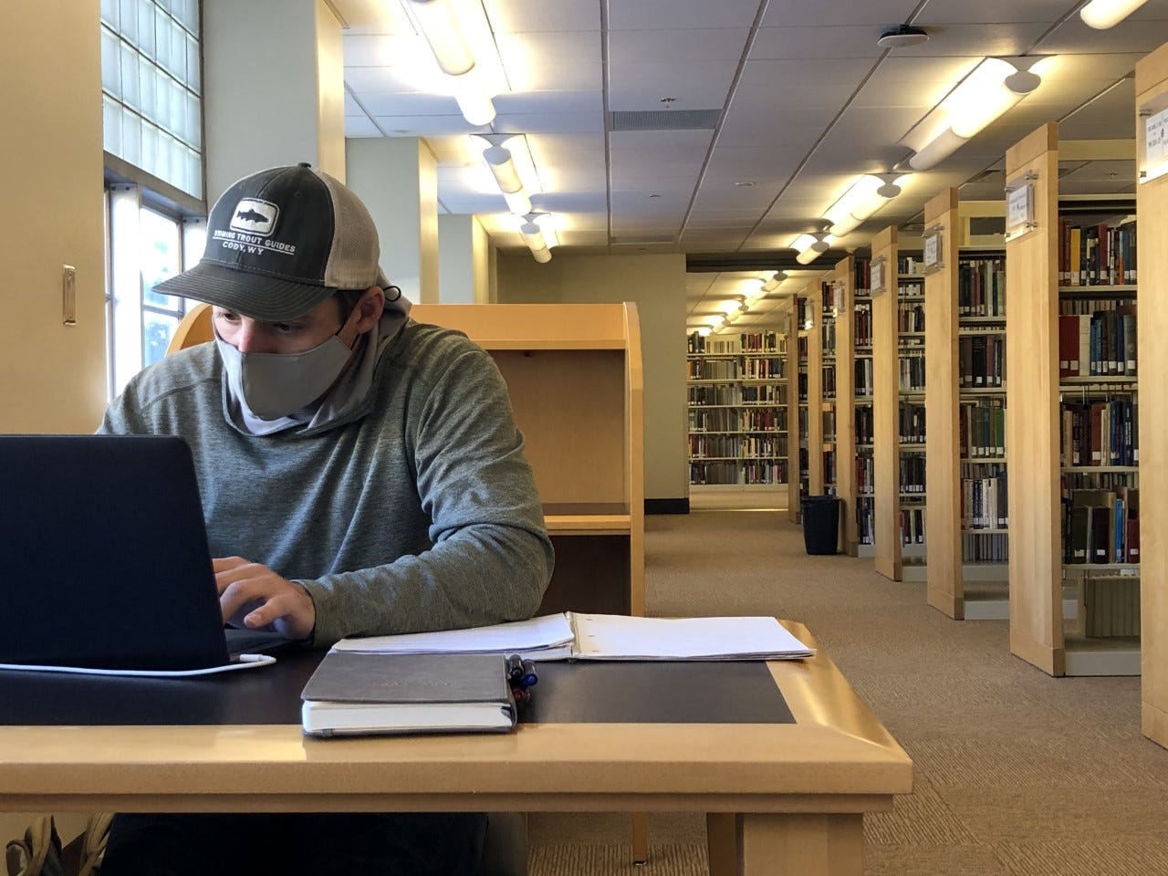 A young man in a gray sweatshirt, ball cap and mask looks at his laptop. Book shelves fade into the background. A young man in a gray sweatshirt, ball cap and mask looks at his laptop. Book shelves fade into the background.