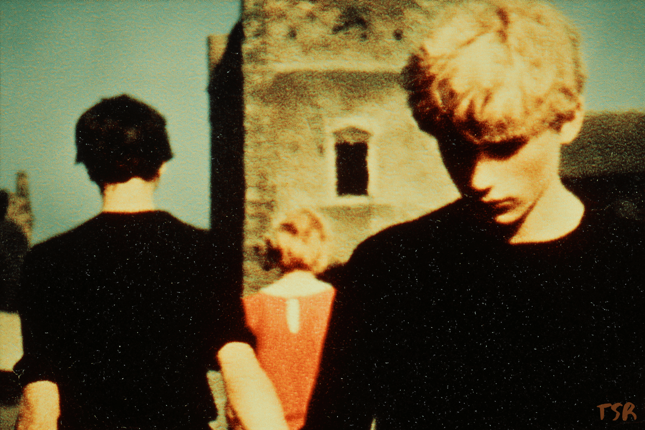 A 1970s-style photograph capturing a blonde teen with a stormy expression, head bowed as he walks away. Behind him, a dark-haired boy moves in the opposite direction, hand reaching back toward him, while a young woman’s figure recedes in the distance.