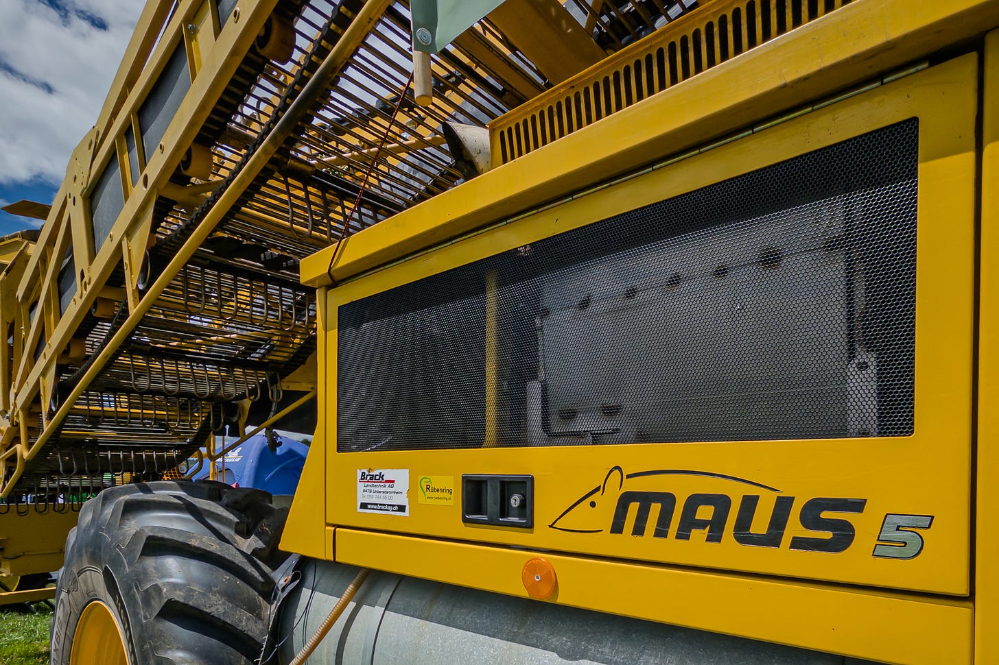 Detailansicht eines gelben Rübenreinigungsladers vom Typ Maus 5. Zu sehen sind das grosse Rad, die seitliche Maschinenverkleidung mit dem Schriftzug „Maus 5“ und darüber die offenen Förder- und Reinigungsgitter der Erntetechnik.