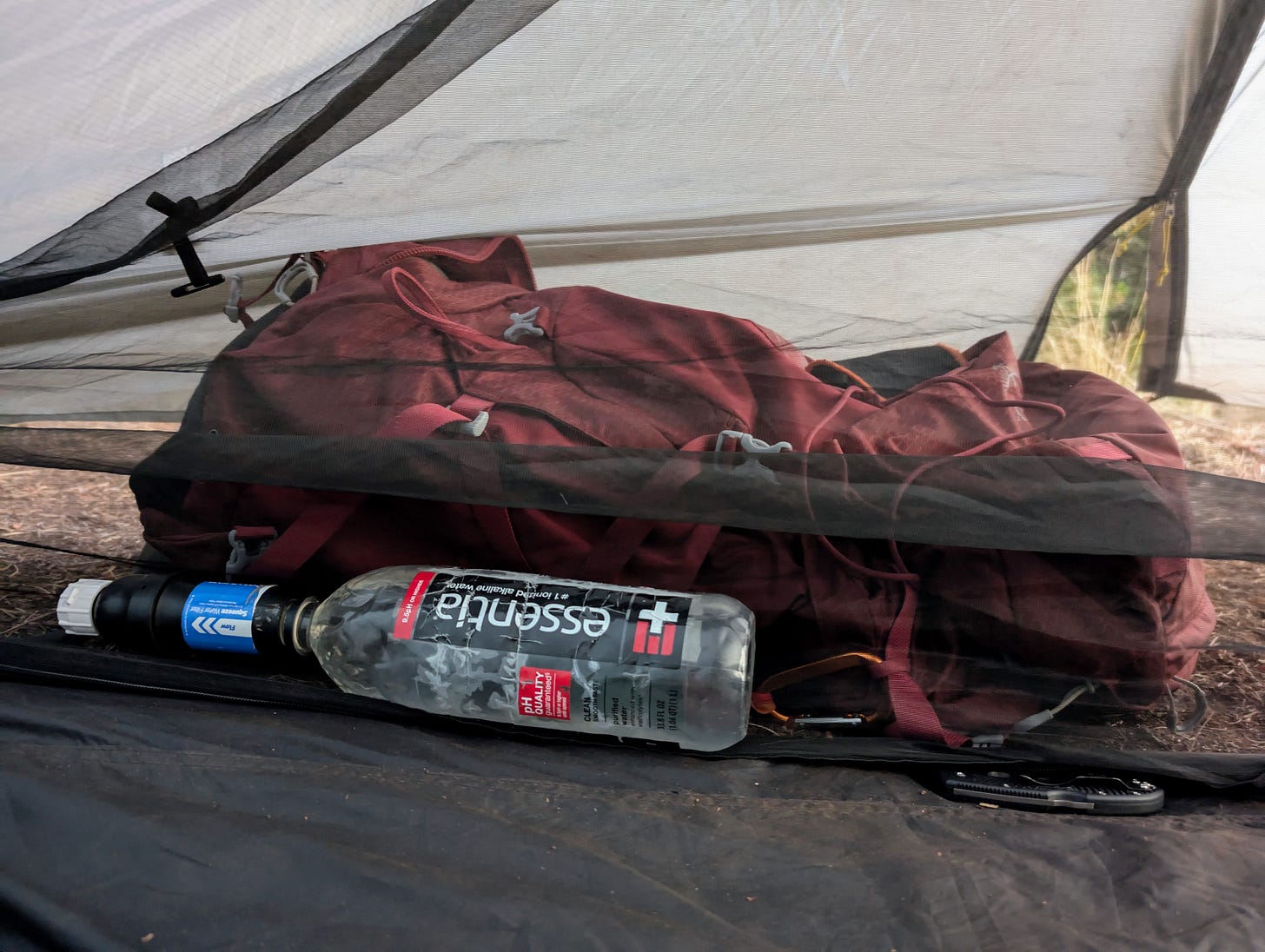Backpack tucked under the tent vestibule beside an Essentia water bottle, resting on the ground under the rainfly. Backpack tucked under the tent vestibule beside an Essentia water bottle, resting on the ground under the rainfly.