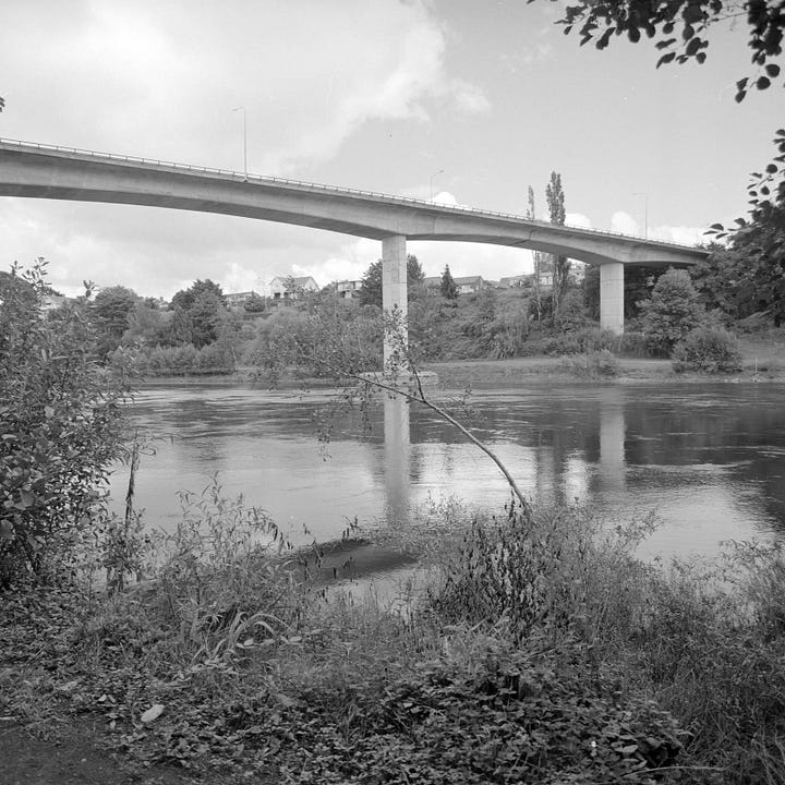 Left image: A screenshot from Google Streetview from on-top of Whitiora Bridge, facing east. It is a three lane bridge, with two lanes going east, and one lane going west, and a small footpath on the south end of the bridge. Right image: A black and white photo of Whitiora Bridge, taken from the north-east side of the river, estimated to be taken in the 1980s.