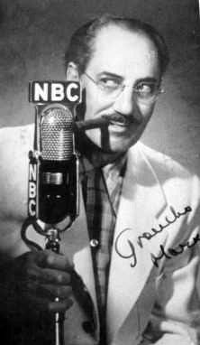 A cigar-chomping, mustauched man stands behind a microphone labeled "NBC."  Picture autographed by Groucho Marx.