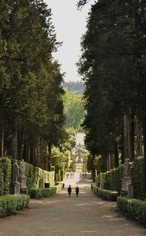This may contain: two people walking down a path in the middle of trees and bushes with statues on either side