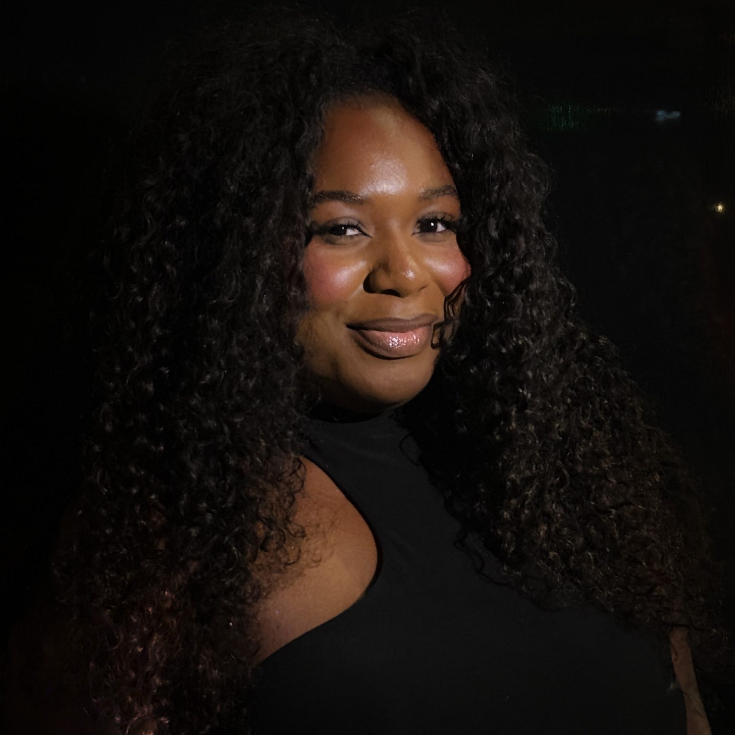 An image of Dejaih Smith, a woman with brown skin and long dark curly hair. She is illuminated by a spotlight standing against a dark background. An image of Dejaih Smith, a woman with brown skin and long dark curly hair. She is illuminated by a spotlight standing against a dark background.