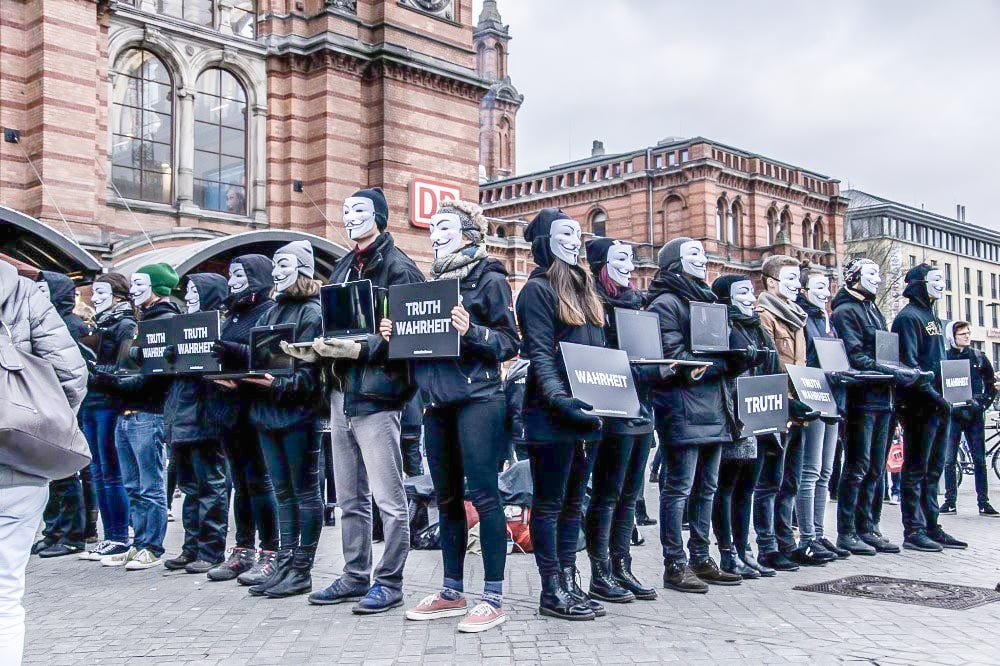 People in black clothes and with Guy Fawkes masks holding up signs and monitors. The signs read "TRUTH | WAHRHEIT". People in black clothes and with Guy Fawkes masks holding up signs and monitors. The signs read "TRUTH | WAHRHEIT".