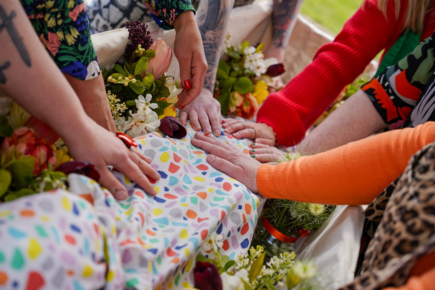 A brightly coloured shroud inside a willow coffin surrounded by jam jars of homegrown flowers. Hands of family gently touch the fully shrouded body.