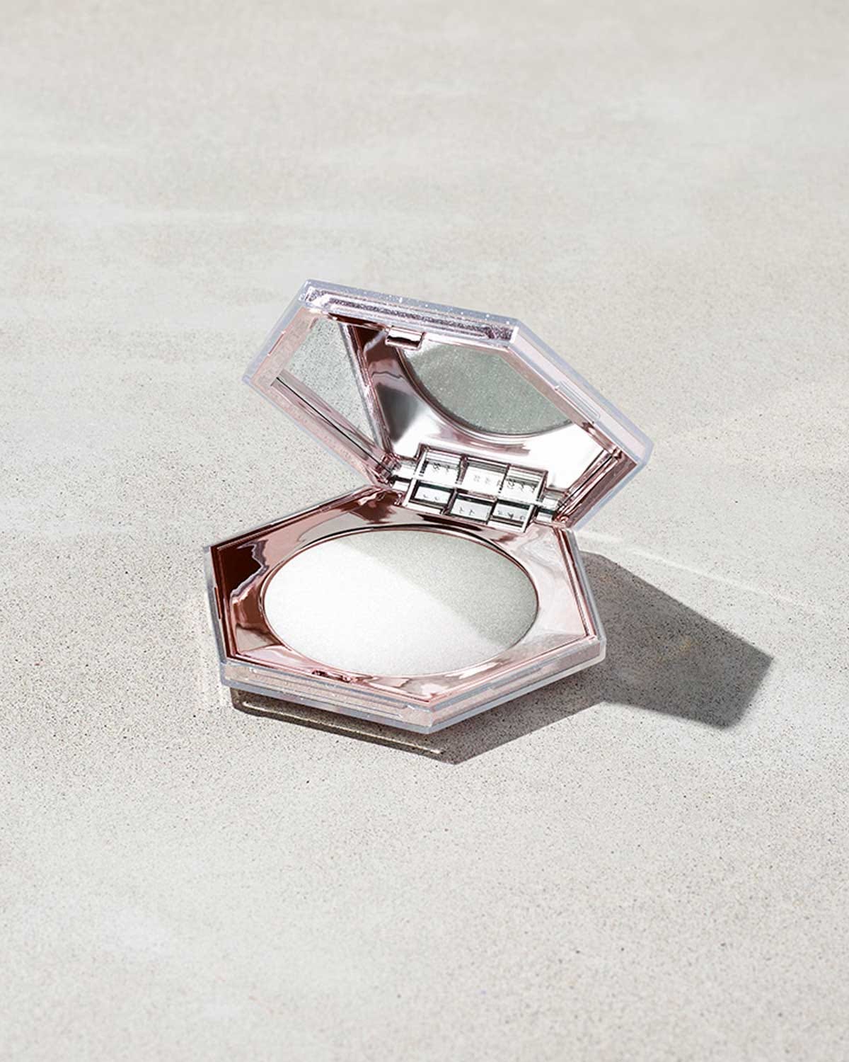 A high-angle product shot of a hexagonal, rose-gold chrome makeup compact opened to reveal a shimmering white-silver crystalline powder. The compact rests on a neutral, textured stone surface with soft shadows.