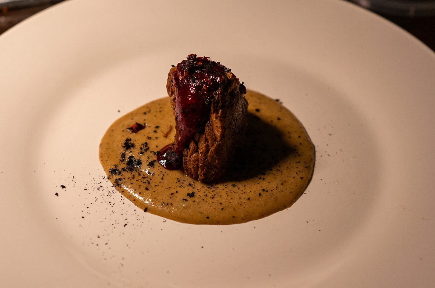A close-up of a plated dish featuring a dense, square cut of braised beef resting in a pool of smooth brown sauce, topped with a dark berry reduction.