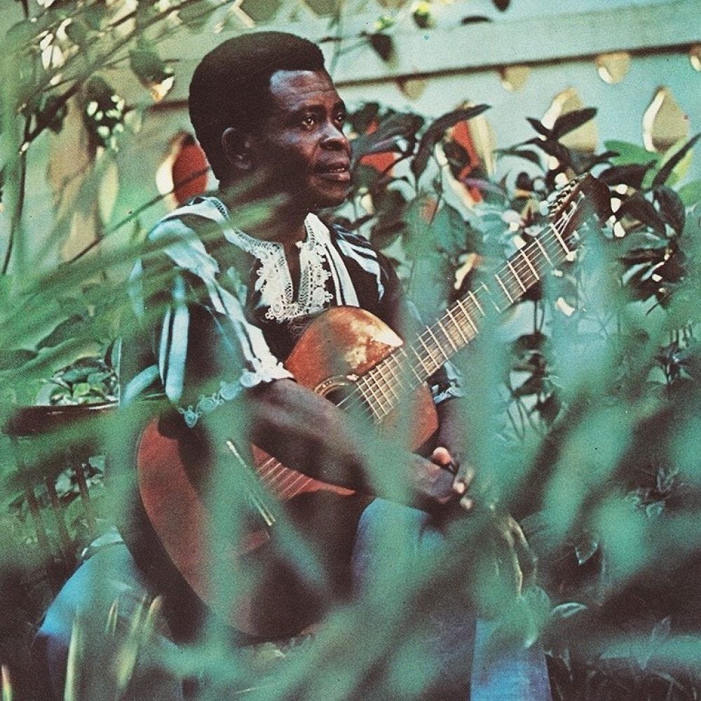An image of Francis Bebey, a black man with a close-cropped afro, maybe in his 40s. He is wearing what looks like a black and white dashiki (West African traditional shirt) and blue jeans. He is looking off-camera, sitting and holding a guitar against his knees and torso, one of his hands wresting against his other wrist. There are green leaves behind and around him, and a white wall of crossing arches beyond. There are blurred leaves in the foreground, obscuring him slightly.