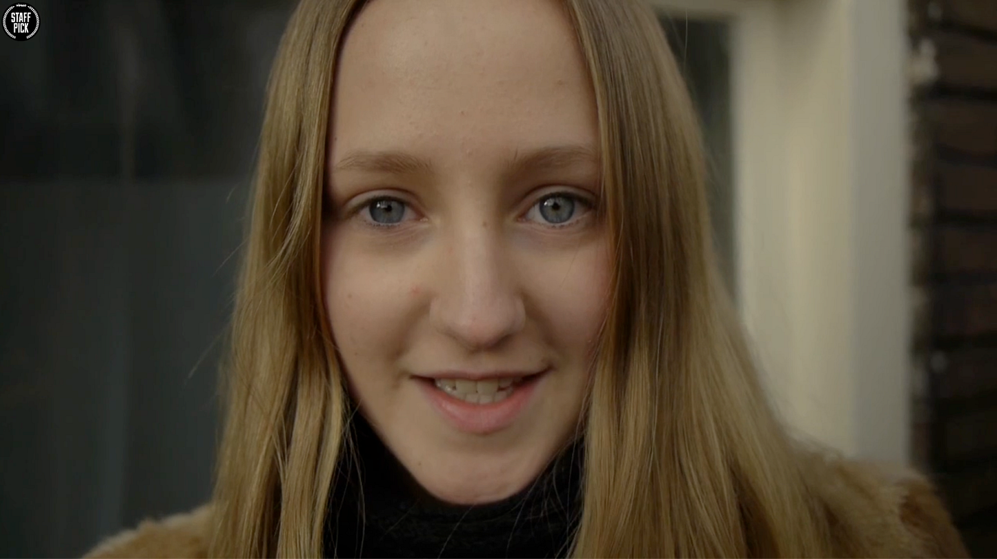 Close-up portrait of a white teenage girl with long straight blonde hair wearing a black turtleneck, smiling gently at the camera indoors.