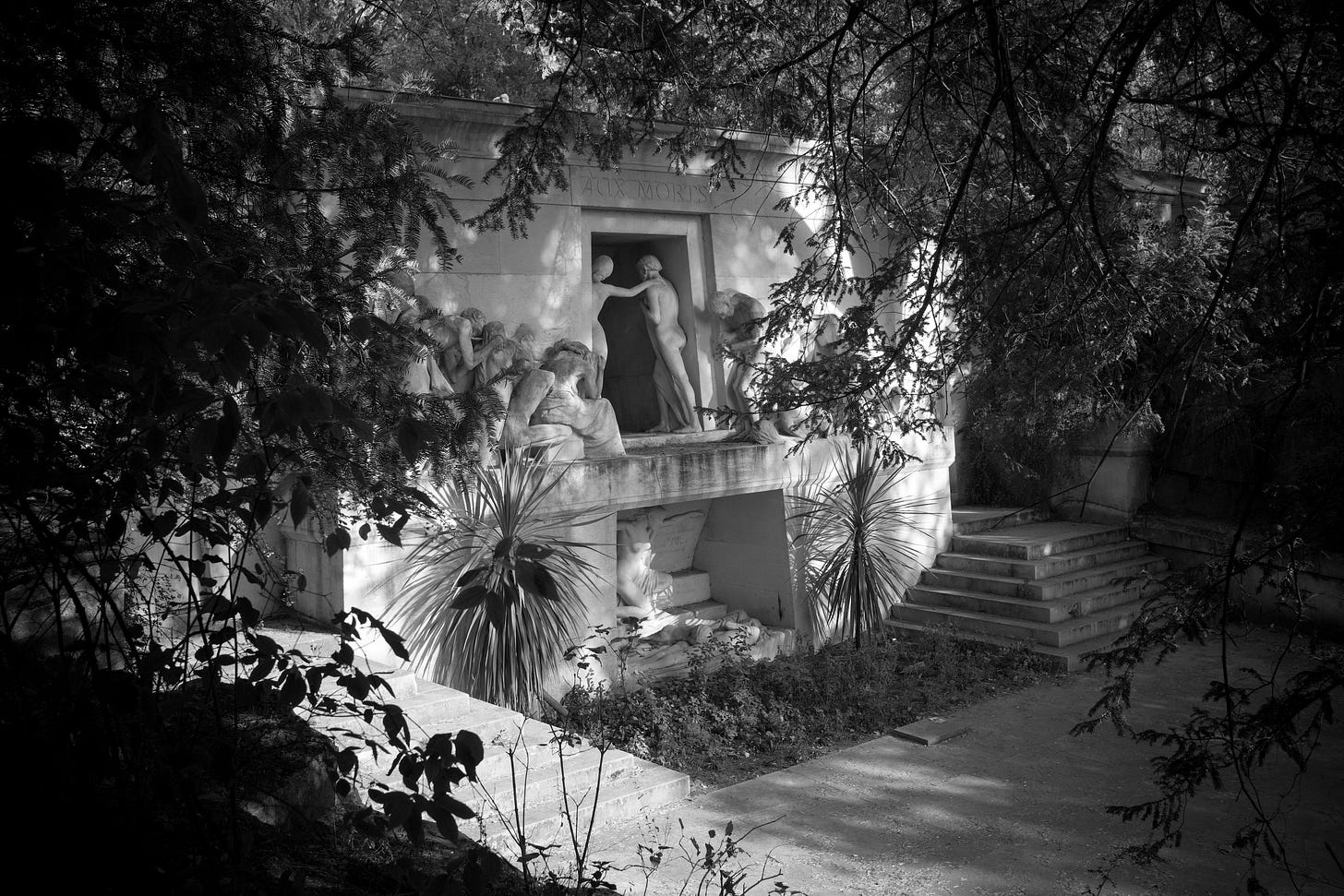 A photo of a large stone monument called, Monument aux Morts. There are carved figures welcoming the dead into a door. It was designed by Paul-Albert Bartholomé in the 19th century, and is a memorial to the unidentified Parisian dead who do not have graves of their own.  Behind the monument is a communal ossuary. The inscription translates to, “For those who live in the shadow of darkness, the light shines.”