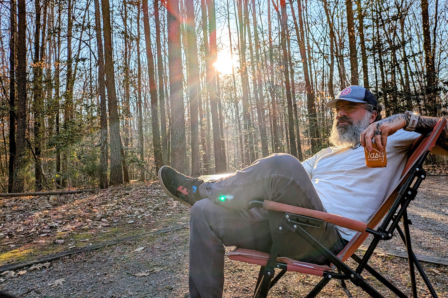 The author reclines in a camp chair in a leafless winter forest, holding a drink in an orange Tasty Beverage Co. koozie. Sunlight filters through bare trees behind him. He wears a trucker cap, glasses, a white t-shirt, and gray pants, with tattooed arms visible.