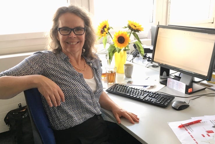 Meredith Wadley sits at an office desk, turning away from her computer monitor to smile at the camera. She wears dark-framed glasses, a blue-polkadot shirt and dark slacks. Various items are on her tidy desk, a glass with tea, a coffee mug, a vase of sunflowers, and a printout of a screenshot of a webpage needing copyediting. Several windows behind her are bleached bright with sunlight.