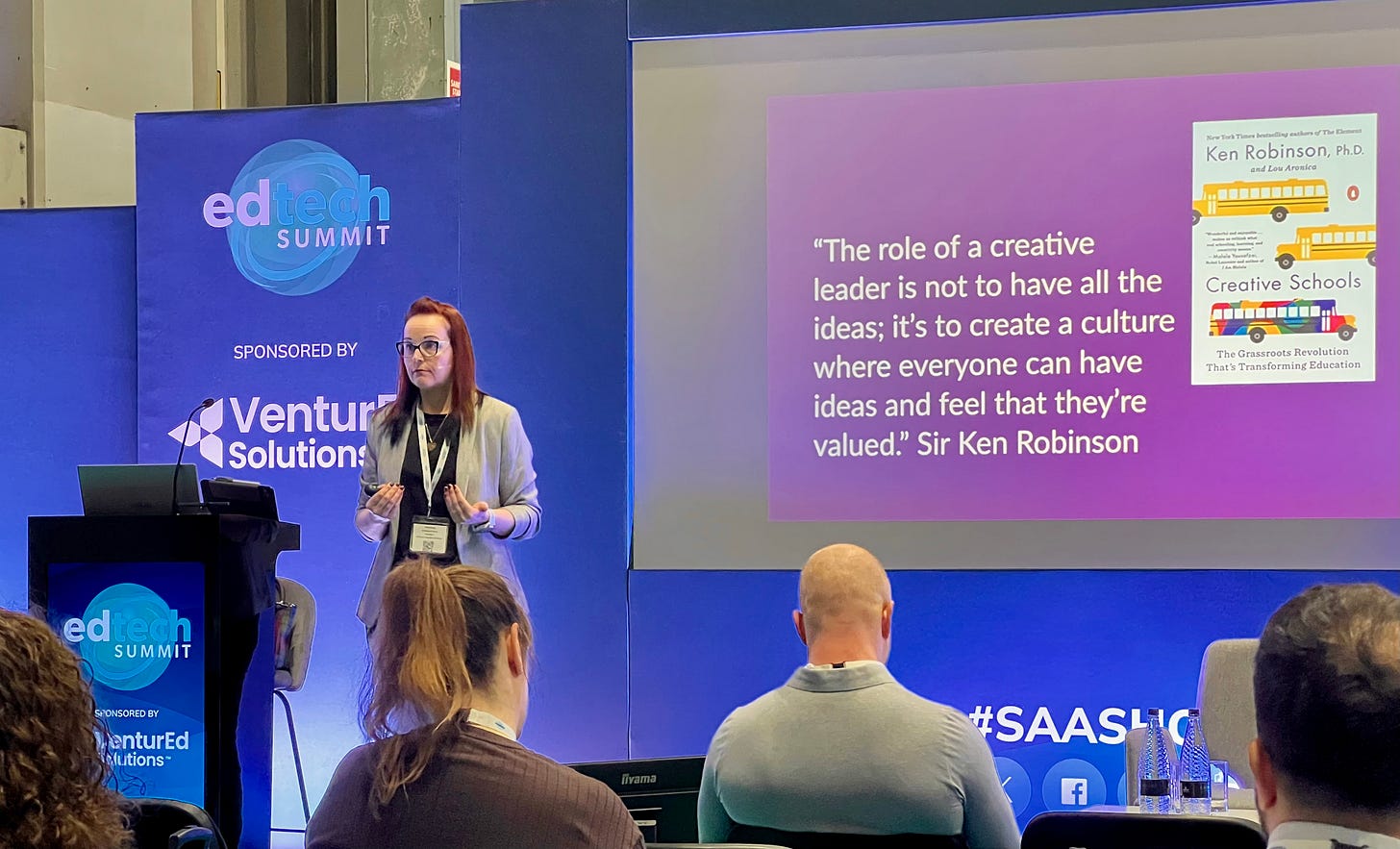 Georgina Dean speaking on stage at the EdTech Summit, engaging the audience with a presentation on visual thinking and the future of work. She is standing confidently at a podium with a microphone, gesturing to emphasize a point. The background features event branding and lighting, creating a professional conference atmosphere.