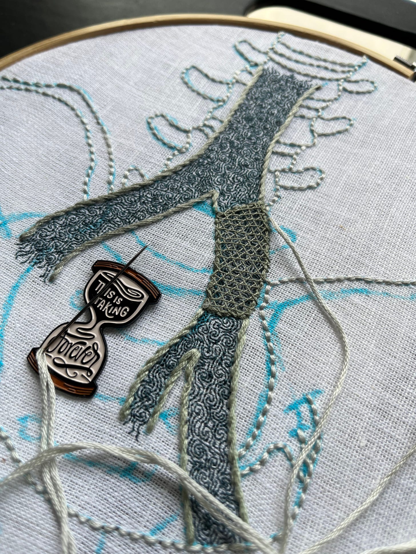 A pelvis, lumbar vertebrae, and part of the iliac vein are sketched in blue marker on white linen. Most of the iliac vein is outlined in grey-green stem stitch and the photo is focused on this area. A section on the left branch is outlined in darker grey-green split stitch with some horizontal and crossing threads stretched across it. Intricate turbulence is stitched in single strand dark aquamarine backstitch within the iliac vein. The bones are being stitched in thick backstitch in a white just slightly bluer than the white linen. An hourglass shaped needle minder reading, “this is taking forever” holds the needle.