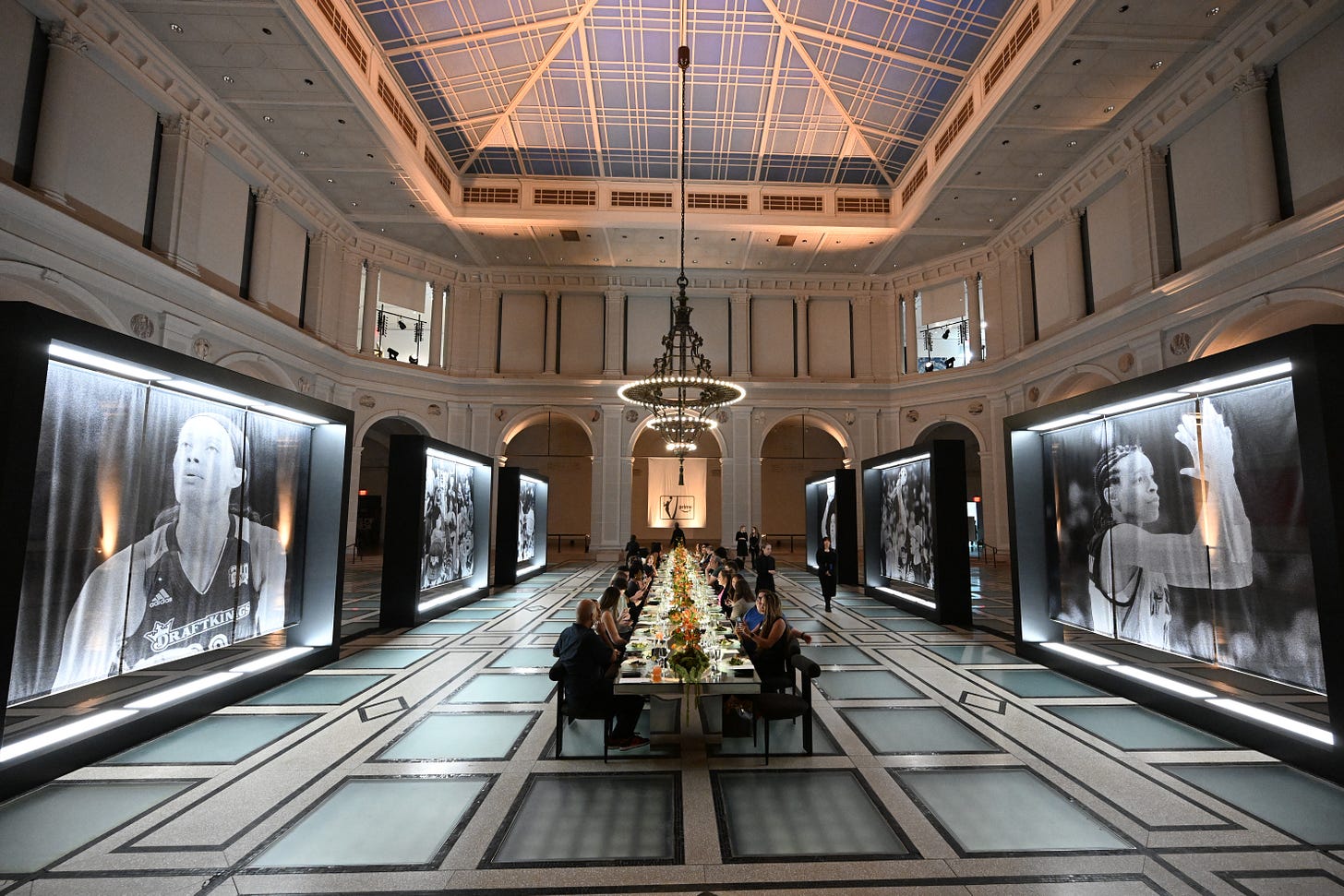 Long dinner table runs through Brooklyn Museum's main hall, flanked by backlit black-and-white portraits of WNBA broadcast talent during their playing days Long dinner table runs through Brooklyn Museum's main hall, flanked by backlit black-and-white portraits of WNBA broadcast talent during their playing days