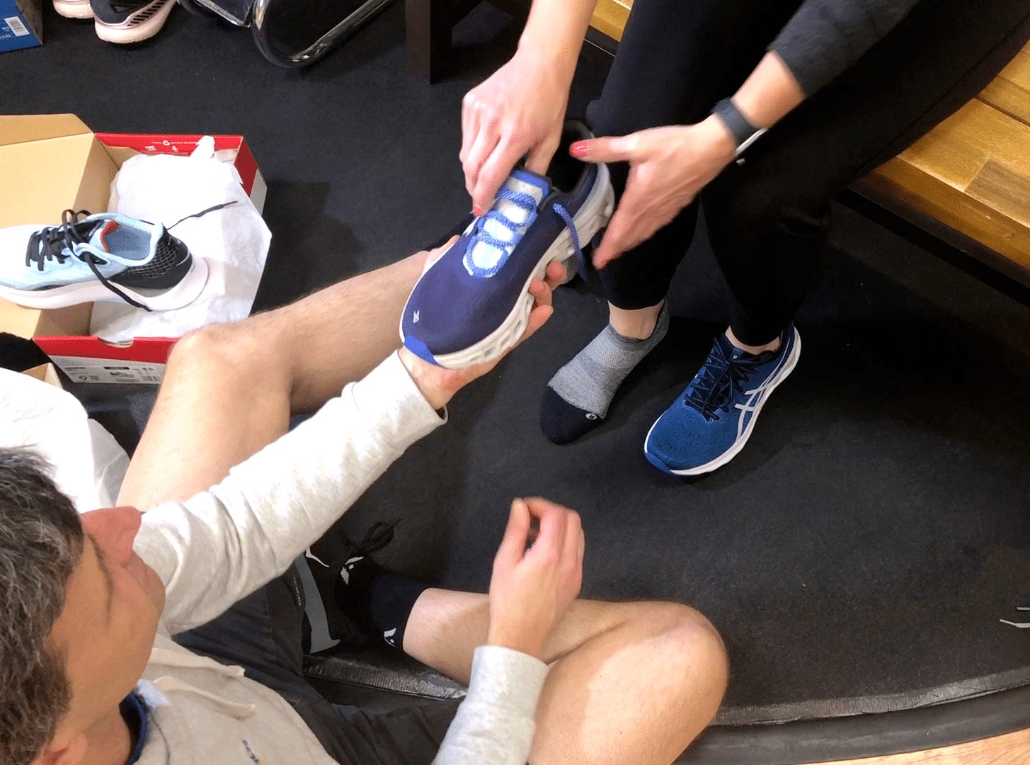 The image shows a person assisting another individual in using a blue, shoe-like device on a black box, surrounded by various shoes and a wooden surface.
AI-generated content may be incorrect.