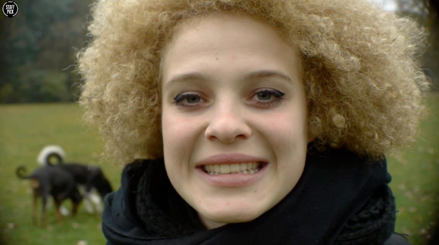 Close-up portrait of a mixed-race teenage girl with curly blonde hair, wearing a black puffy jacket, smiling at the camera outdoors.