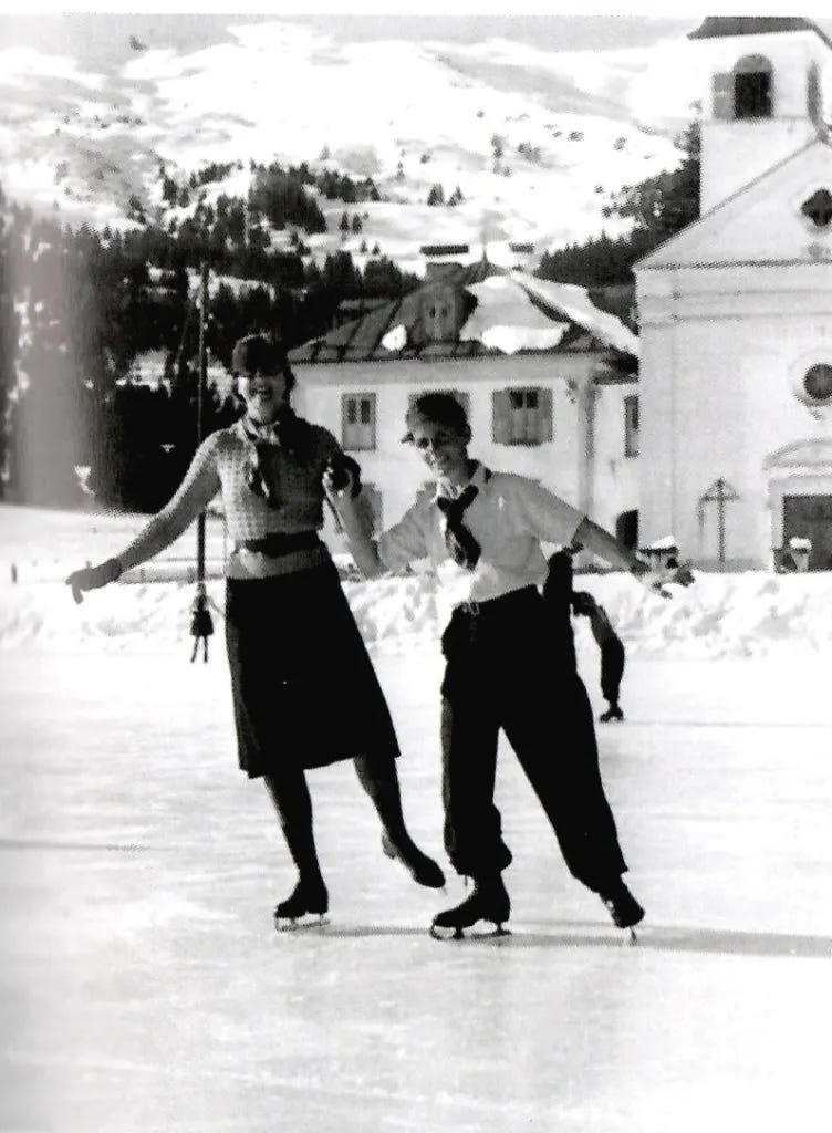 Vintage black-and-white photo of two smiling people ice skating hand-in-hand on a snowy outdoor rink, with a church and snowy hills in the background. The person on the left is wearing a blouse with a scarf and belt, and a skirt, while the person on the right is wearing a button-down shirt, tie, and pants. They are both looking at the camera. Vintage black-and-white photo of two smiling people ice skating hand-in-hand on a snowy outdoor rink, with a church and snowy hills in the background. The person on the left is wearing a blouse with a scarf and belt, and a skirt, while the person on the right is wearing a button-down shirt, tie, and pants. They are both looking at the camera.