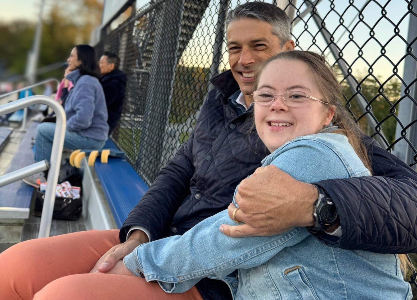 Peter and Penny sit together on metal bleachers, smiling warmly at the camera. Peter has his arm wrapped around Penny. They are both wearing jackets. A chain-link fence and other spectators are visible in the background.