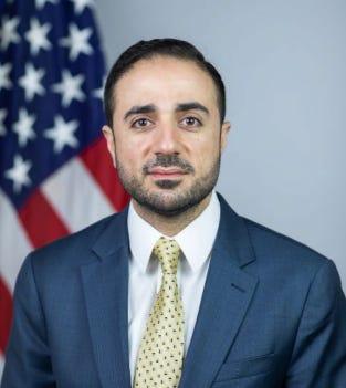 First image shows a middle-aged man with short dark hair and mustache wearing a dark blue suit white shirt and yellow tie with polka dots standing in front of two American flags against a light gray background in a formal setting. Second image depicts a man with short dark hair and mustache dressed in black long-sleeve shirt black pants and white belt wearing a red and white keffiyeh headscarf raising his right arm upward with palm open in a room with beige walls and abstract colorful paintings on the walls including red orange and multicolored striped artwork.