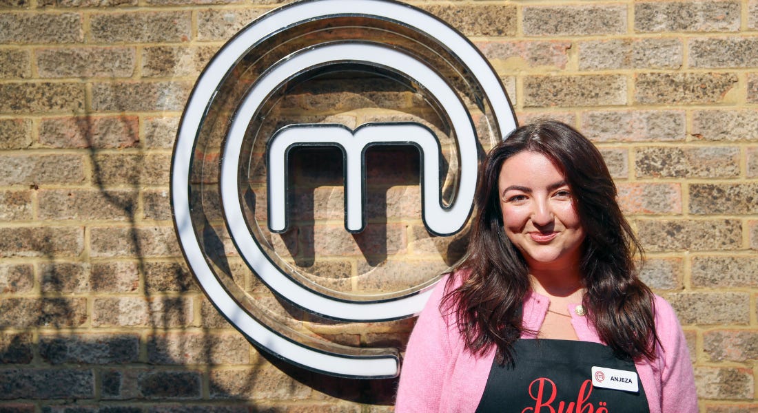 A woman - Anjeza Popaj - wearing a pink shirt and apron standing in front of a brick wall with the Masterchef logo on it
