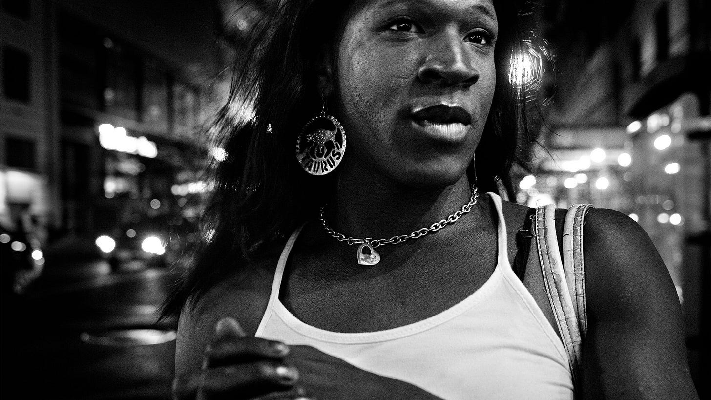 A high-contrast black-and-white photograph of a Black woman wearing a white tank top, a thick chain necklace and big Taurus earrings. She looks off camera while standing on a city street at night.