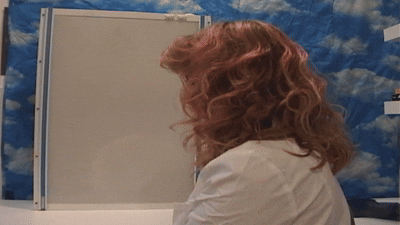 Zsofi dressed as a doctor in a lab coat, snapping on pink exam gloves. Behind her is a vintage x-ray viewer light box and a cloud print tapestry.