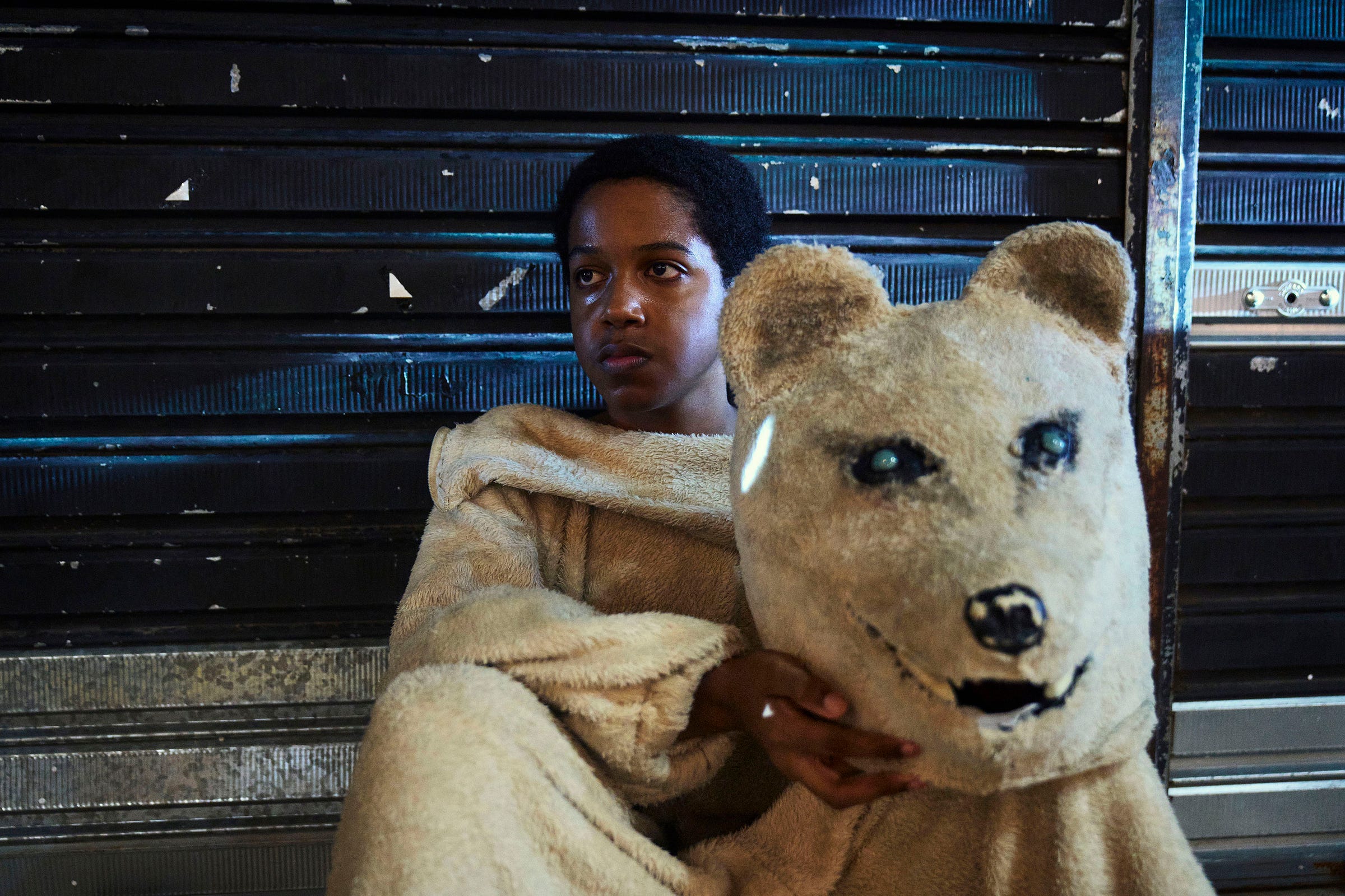 A person in a light brown bear costume sits against a dark metal shutter, holding the bear head in their lap and looking to the side with a thoughtful expression.