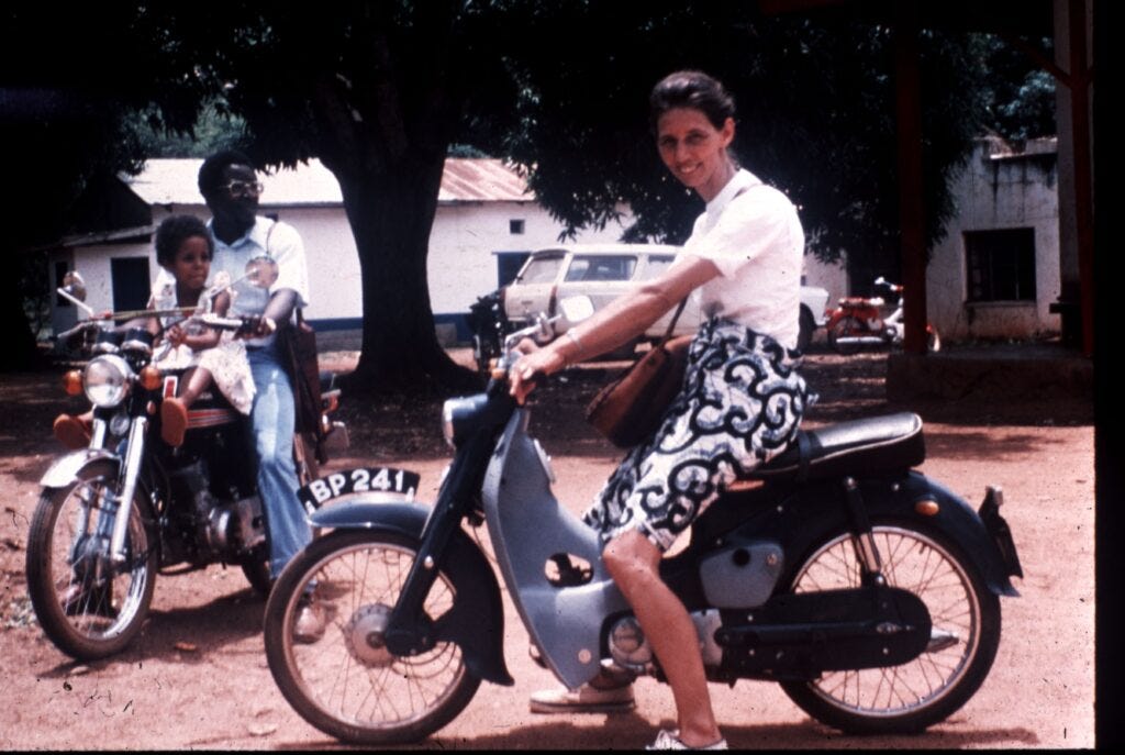 A woman smiles as she sits on a motorbike in the foreground, while in the background, a man rides a motorcycle with a young child sitting in front of him. They are outdoors on a dirt road, with trees and buildings in the background. A woman smiles as she sits on a motorbike in the foreground, while in the background, a man rides a motorcycle with a young child sitting in front of him. They are outdoors on a dirt road, with trees and buildings in the background.