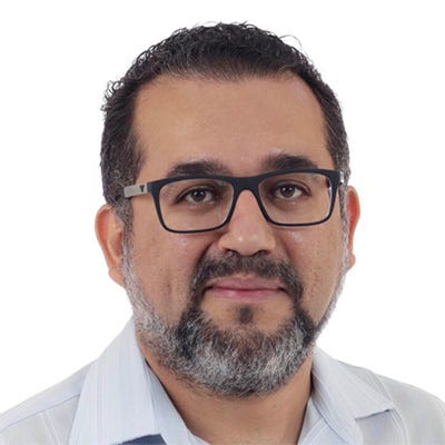 Homem com cabelo curto e escuro, barba grisalha, um leve sorriso e óculos. Veste uma camisa social branca. Homem com cabelo curto e escuro, barba grisalha, um leve sorriso e óculos. Veste uma camisa social branca.