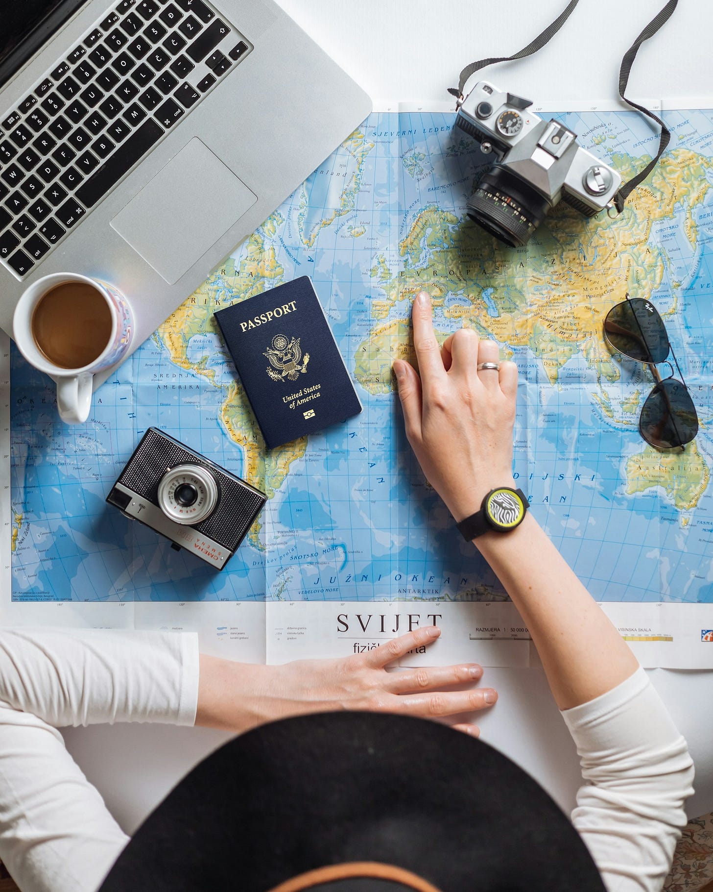 Mappa del mondo aperta su un tavolo. La mano di una persona bianca ripresa dall'alto indica un punto sulla mappa. Poggiati sulla mappa ci sono un passaporto statunitense, un laptop, due macchine fotografiche, occhiali da sole e una tazza di caffè.