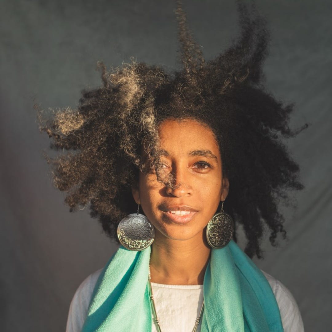 Photo de Alexis Pauline Gumbs, une femme Noire avec ses cheveux crépus qui encadrent son visage, habillée d'une écharpe vert clair et de grosses boucles d'oreilles rondes