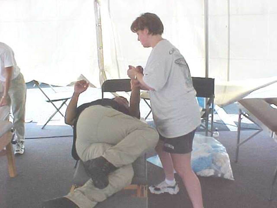 Chiropractor in a white T-shirt adjusts the neck of a man seated in a chair inside a white tent