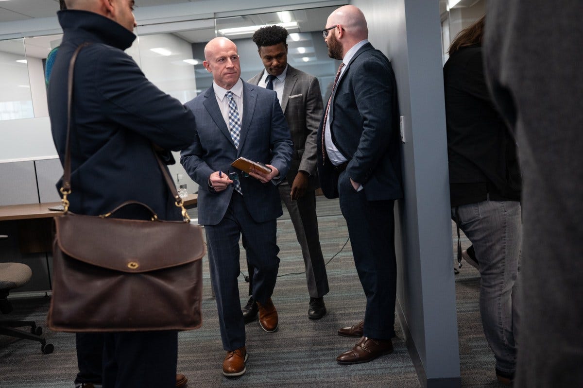 Four men in business attire stand and talk in an office hallway; one holds a notepad and pen, and another holds a phone.