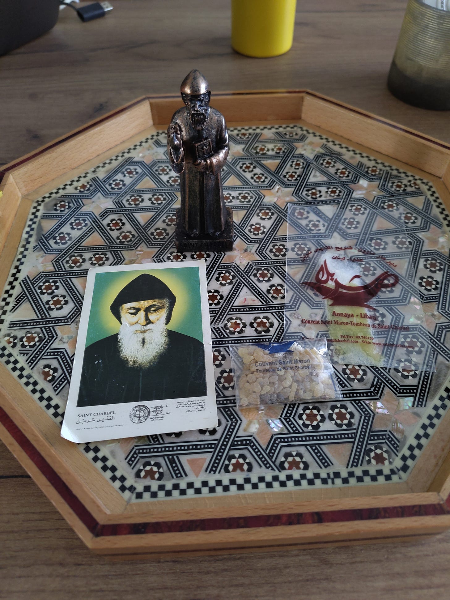 A statue and picture of Saint Charbel on a hexagonal tray, along with a packet of incense and a packet of oil.
