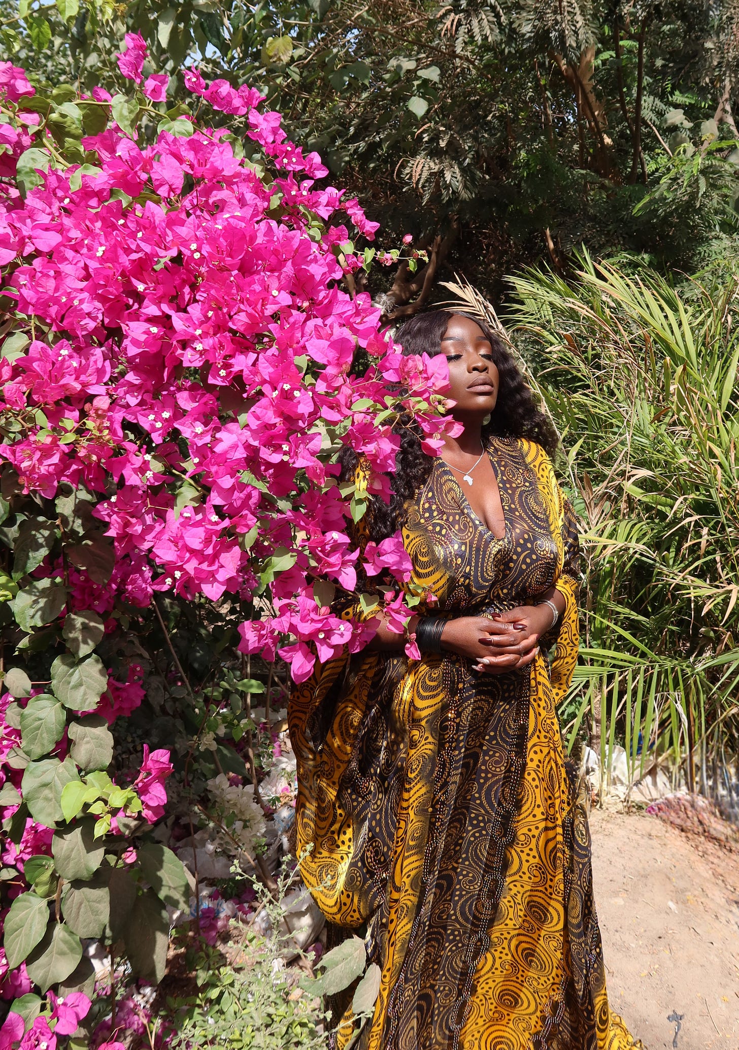 Lovette Jallow standing in Gambia beneath tall banana trees, wearing a gold and black traditional patterned dress, embodying African wisdom and storytelling traditions. Lovette Jallow standing in Gambia beneath tall banana trees, wearing a gold and black traditional patterned dress, embodying African wisdom and storytelling traditions.