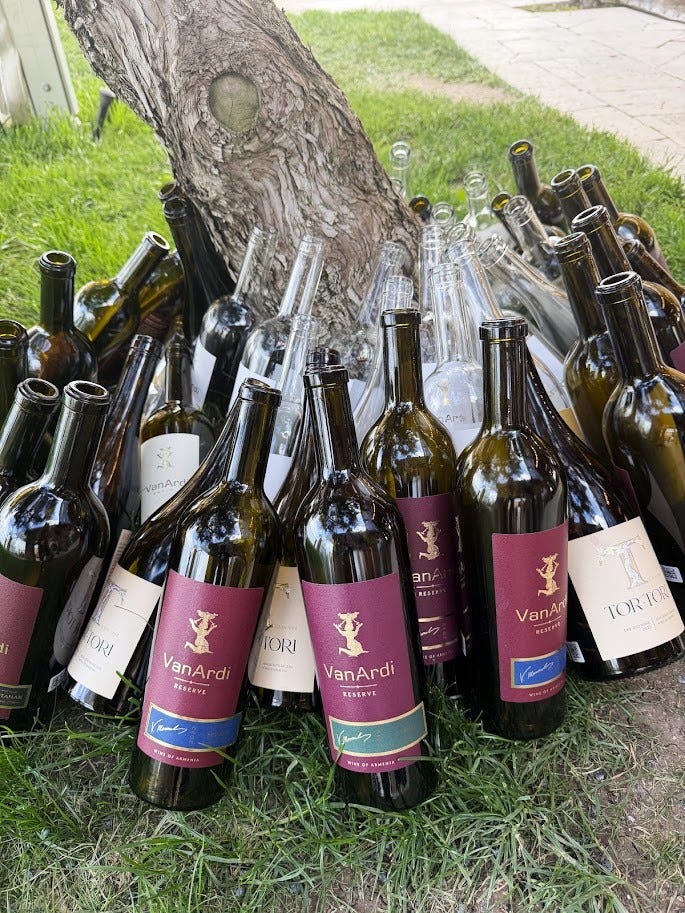 Van Ardi's stone estate building and belltower overlooking green hillside gardens, alongside a collection of empty Van Ardi and Tor-Tori wine bottles gathered under a tree after tasting