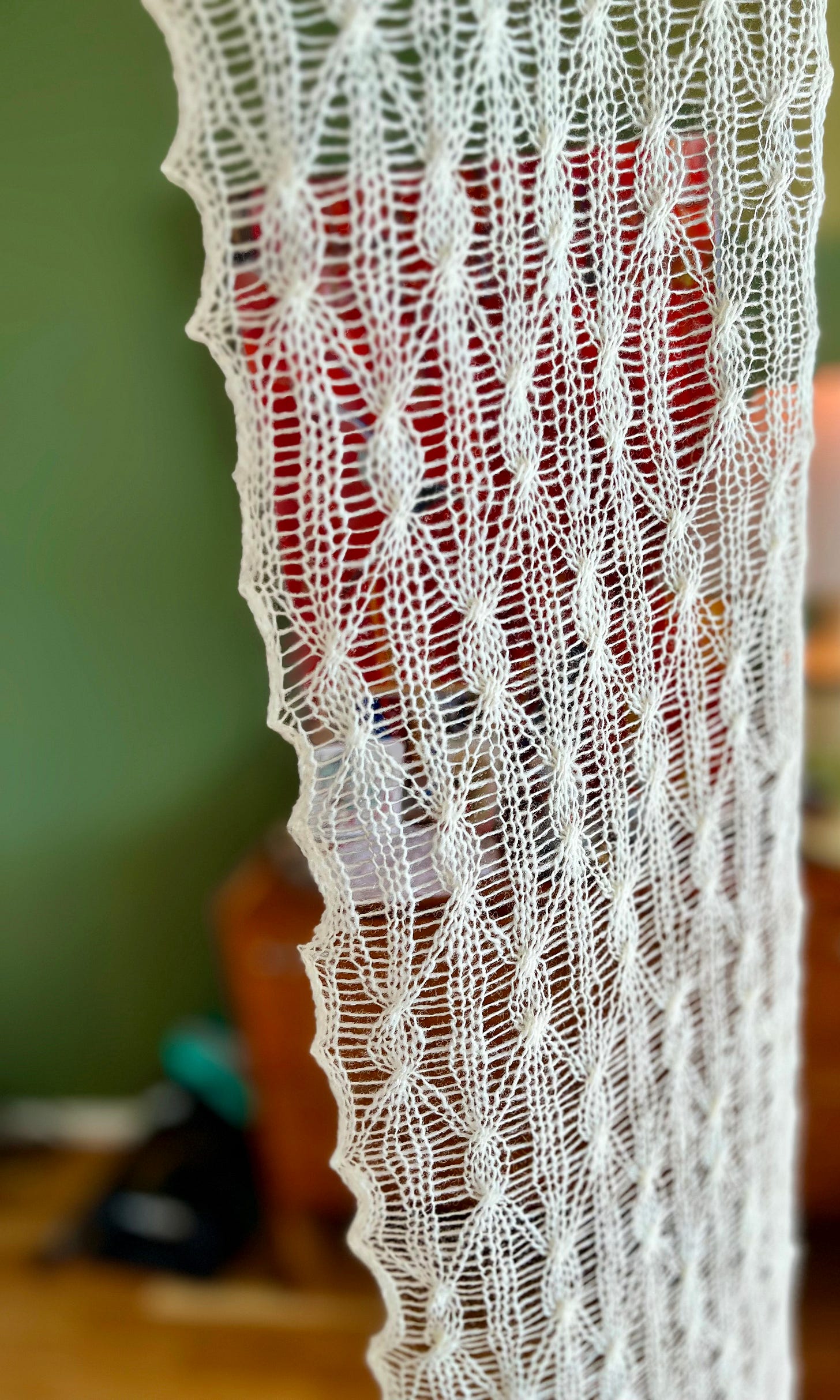 A finished cream lace scarf hanging vertically against a sage green wall. The full length is visible, showing the repeating elongated diamond pattern, scalloped side edges, and the fine drape of the single-ply wool.