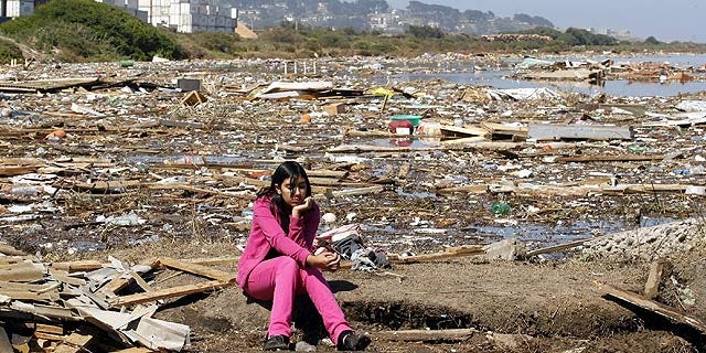 menina sentada pensativa entre os escombros do terremoto em uma praia da cidade de concepción, no chile. ela tem os cabelos pretos e uma um conjunto rosa.