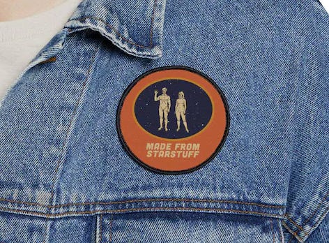 A circular embroidered patch displayed on a denim jacket. The patch features the nude human figures from the 1972 Pioneer plaque—one male, one female—standing in front of a dark starry background. Below them, in bold lettering, is the phrase “MADE FROM STARSTUFF,” a reference to Carl Sagan’s famous quote about our cosmic origins. The border of the patch is orange with a black edge. A circular embroidered patch displayed on a denim jacket. The patch features the nude human figures from the 1972 Pioneer plaque—one male, one female—standing in front of a dark starry background. Below them, in bold lettering, is the phrase “MADE FROM STARSTUFF,” a reference to Carl Sagan’s famous quote about our cosmic origins. The border of the patch is orange with a black edge.