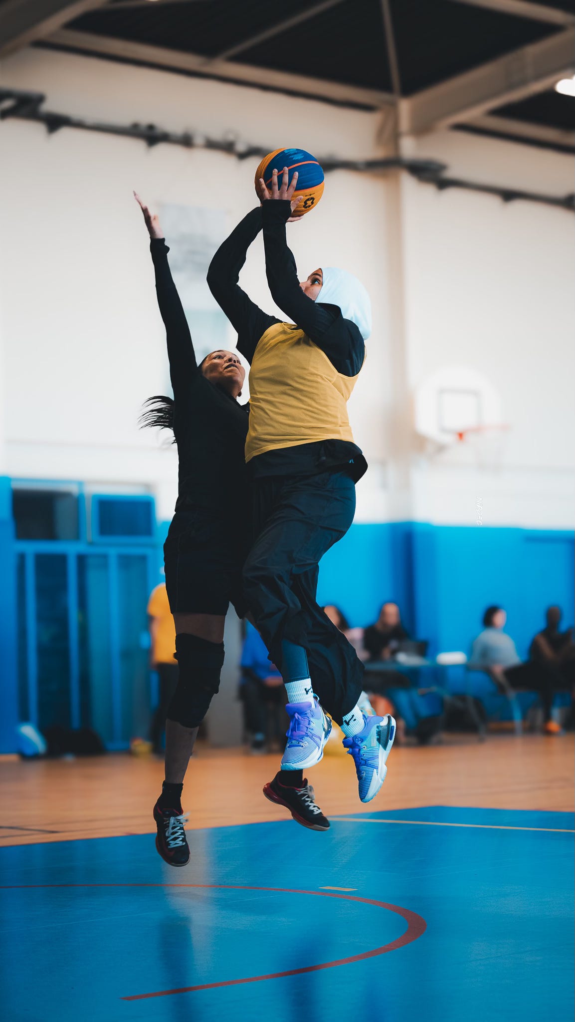 Tournoi 3x3 au club de basket-ball de Noisy-le-Sec, le 13 avril 2024. Une basketteuse portant un couvre-chef sportif vient de faire un saut. Elle est dans les airs et s'apprête à tirer le ballon de basket. Une autre basketteuse tente de l'en empêcher pour défendre son équipe.